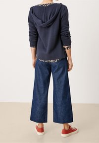 Nainen, jolla on yllään tummansininen huppari, kukkakuosiset paita, leveät tummat farkut ja punaiset kangaskengät, kääntyy poispäin yksinkertaisella taustalla.