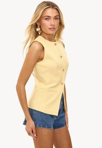Geel mouwloos vest met een getailleerde pasvorm, voorzien van vijf knopen in goudkleur en zijzakken, gecombineerd met denim shorts. Glad textuur.