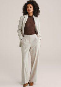 Naine seisab käed taskutes, kandes helebeezhi ülikonda, pruunikat tekstuuriga topi ja pruune kontsakingi, taust on lihtne.