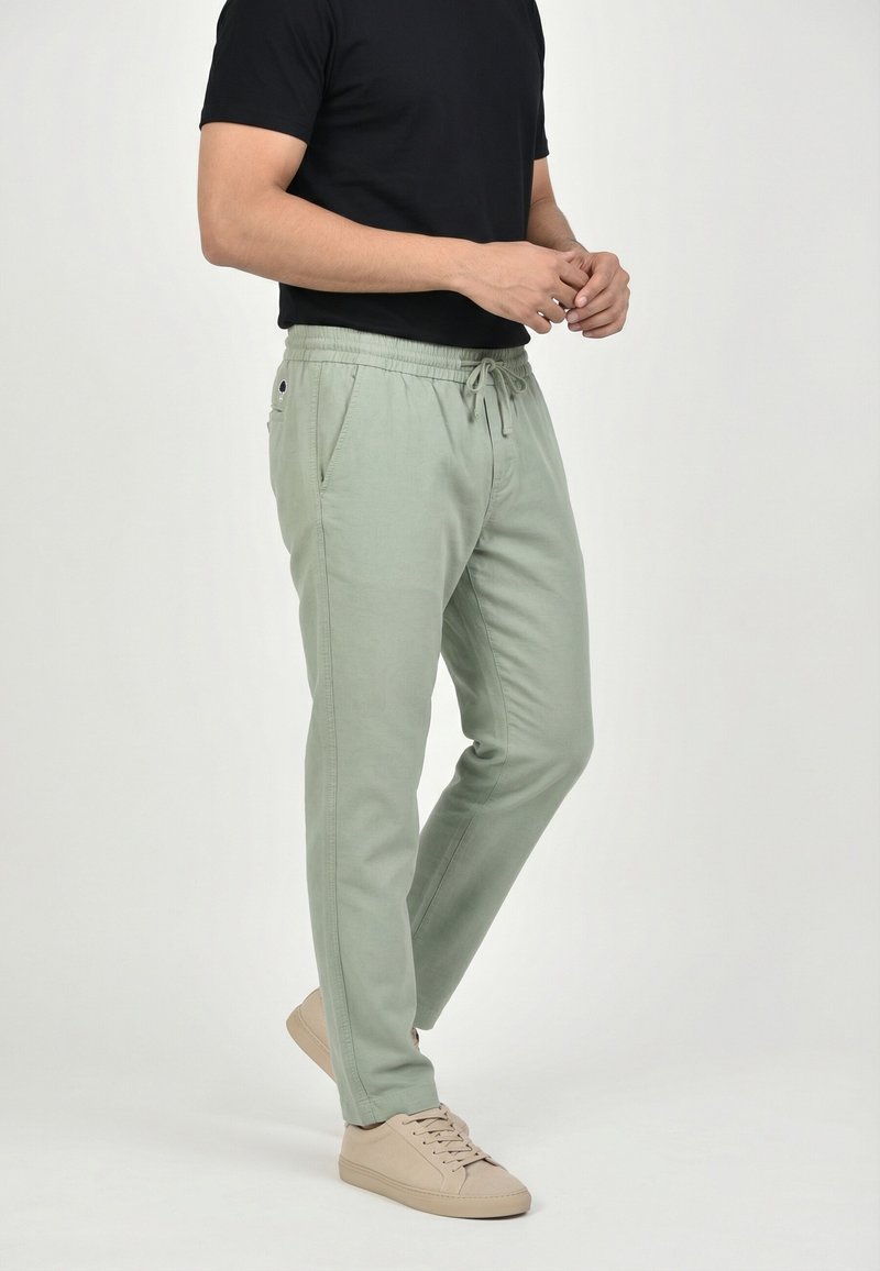 Uomo che indossa pantaloni verdi chiaro con coulisse, maglietta nera a maniche corte e scarpe da ginnastica beige, in piedi contro uno sfondo semplice.