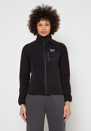 Frau mit lockigem Haar, die eine schwarze Reebok Fleecejacke mit Reißverschluss und graue Hosen vor einem schlichten hellen Hintergrund trägt.
