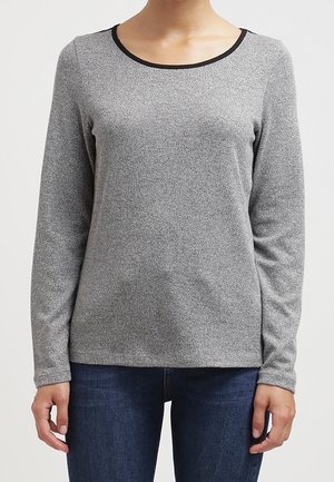 Kobieta w dopasowanej szarej bluzce z długim rękawem z czarnym wykończeniem przy okrągłym dekolcie i ciemnych niebieskich dżinsach, stojąca na jednolitym tle.