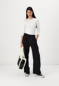 Μαύρα παντελόνια σε φαρδιά γραμμή με πιέτες μπροστά, συνδυασμένα με ένα εφαρμοστό λευκό μπλουζάκι μακρυμάνικο και μια μαύρη τσάντα από ψεύτικο δέρμα με λευκές λεπτομέρειες.