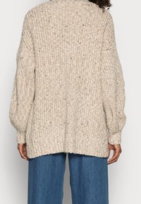 Beige stickad cardigan med ett texturerat mönster, nedhängda axlar och en något längre bakslut, kombinerad med vida blå jeans.