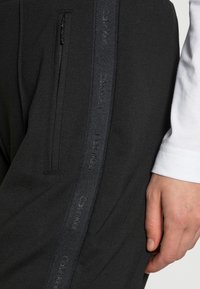 Zwarte sweatpants met een zijzak met ritssluiting, voorzien van een gestructureerde zwarte streep met reliëf 'Calvin Klein' merknaam langs het been.