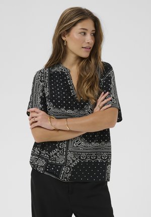 Femeie cu păr lung, purtând o bluză neagră cu mâneci scurte imprimată, pantaloni negri și bijuterii aurii, privind spre lateral cu brațele încrucișate.