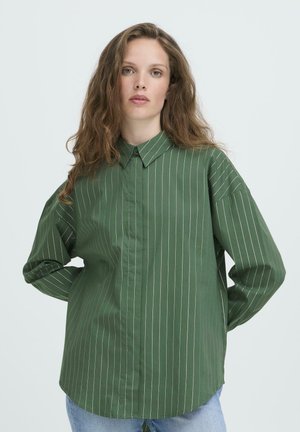 Woman with long wavy hair wearing a green pinstripe button-up shirt and light blue jeans, standing with hands behind her back.
