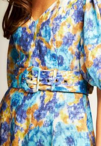 Blau- und gelb gemustertes Kleid mit V-Ausschnitt, kurzen Ärmeln und einem breiten Gürtel mit einer runden Schnalle und metallischen Ösen.