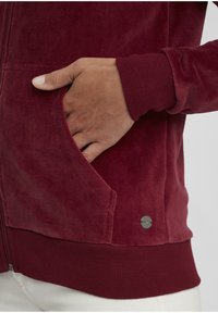 Burgunderfarget zip-jakke med ribbestrikkede mansjetter og nederkant, med et mykt tekstilmateriale og sidelomme med metallakenter.