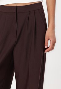 Pantalonii burgundy au un material neted, un design plisat și un tiv. O mână este plasată lângă pantaloni pentru a evidenția detaliile.