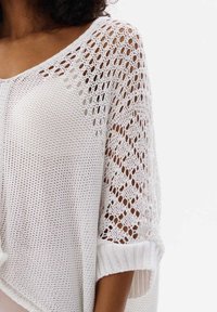 Weißer Strickpullover mit offenem Strickmuster, lockerem Schnitt und aufgekrempelten Ärmeln. Die strukturierte Stoffqualität unterstreicht das atmungsaktive Design.