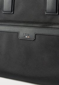 Μαύρη υφασμάτινη τσάντα χειρός με δερμάτινες λεπτομέρειες, διαθέτει κλείσιμο με φερμουάρ και πλακέτα με λογότυπο. Συμπαγής σχεδιασμός με ενισχυμένες λαβές.