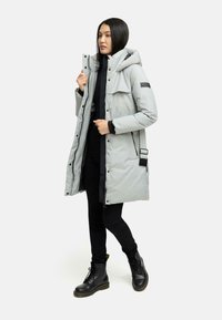 Lys grå isoleret parka med hætte, forreste lommer og sorte lynlåsdetaljer, båret over et sort outfit og parret med sorte kampstøvler.