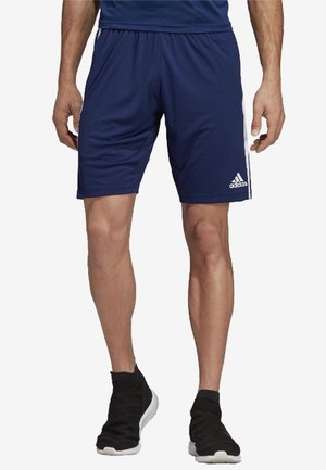 Mężczyzna ubrany w granatowe sportowe szorty Adidas z białymi paskami i czarne wsuwane tenisówki, stojący na białym tle.