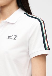 Λευκό πόλο μπλουζάκι με κεντημένο λογότυπο EA7. Διαθέτει μπλε σκούρες ρίγες στους ώμους με πράσινες, λευκές και κόκκινες λεπτομέρειες, καθώς και υφή στις ρίγες.