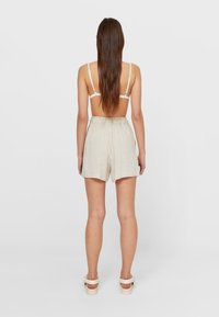 Vrouw staat met de rug naar ons toe, draagt een witte beha, beige hoge taille shorts en witte sandalen met banden tegen een effen achtergrond.