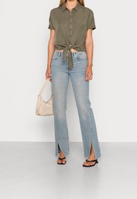 Maglietta verde oliva a maniche corte con nodo frontale, jeans flare blu chiaro con fessure laterali e sandali neri. Tiene in mano una borsa beige.