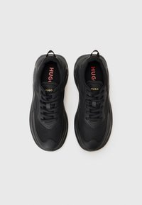 Black sneakers with a textured fabric upper, round toe, and lace-up closure. Features a red and gold logo on the tongue. Thick rubber sole.