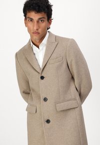 Beige Wollmantel mit maßgeschneidertem Design; verfügt über einen Reverskragen, drei schwarze Knöpfe und zwei vordere Taschen. Glatte Textur, mittellang.