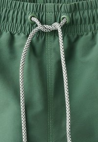 Zelený elastický pás s bielou a čiernou vzorovanou šnúrkou, zaviazanou na uzol na ľahkej tkanine, s viditeľným stredovým prešitím.