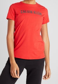 Rød bomulds T-shirt med korte ærmer og rund halsudskæring, med et sort "CALVIN KLEIN" logo over brystet. Glat tekstur.