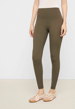 Kvinde iført olivengrønne leggings med høj talje og lyserøde balletflade sko, stående mod en ensartet lys baggrund.