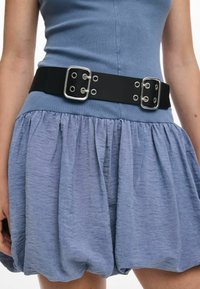 Centură neagră cu accesorii din argint și ochiuri, purtată peste o fustă texturată, de culoare albastru deschis, cu plisuri. Centura are o cataramă rectangulară.