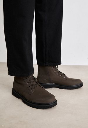 Brązowe skórzane botki za kostkę z okrągłymi noskami, metalowymi oczkami i czarnymi gumowymi podeszwami, w połączeniu z czarnymi dopasowanymi spodniami. Teksturowane wykończenie.