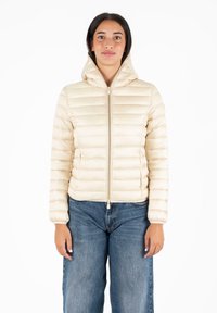 Beige pufferjack met een capuchon, doorgestikt design, vooraan met ritssluiting en elastische manchetten. Draagt over baggy blauwe jeans.