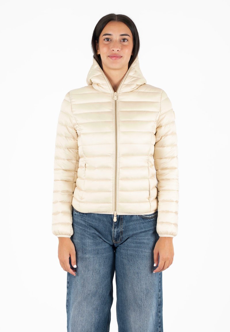 Beige pufferjack met een capuchon, doorgestikt design, vooraan met ritssluiting en elastische manchetten. Draagt over baggy blauwe jeans.