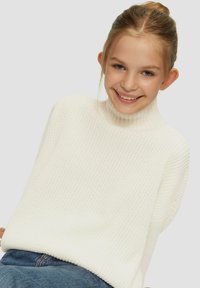 Pull en tricot blanc à col haut, avec une coupe décontractée et un motif texturé. Associé à un jean bleu, sur fond neutre.