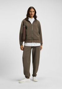 Brun zip-up hoodie med mjuk textur, lager på lager över en vit skjorta. Matchas med bruna avsmalnande byxor och vita sneakers.