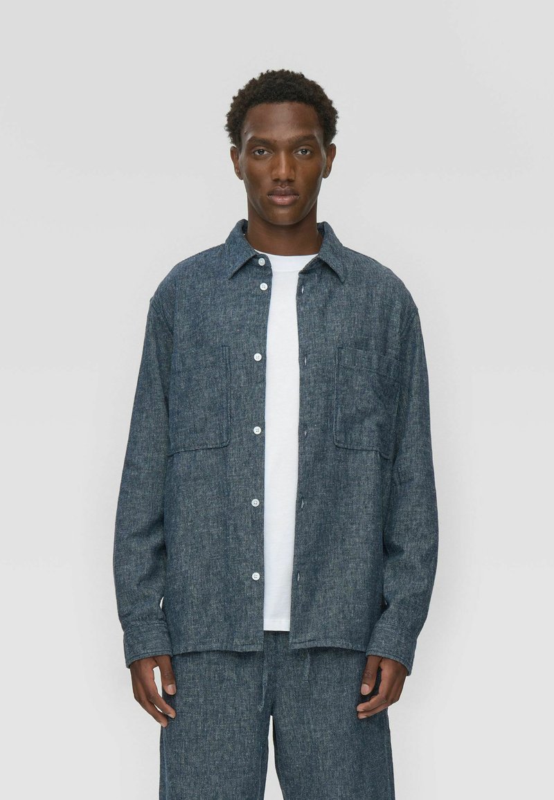 Chemise en denim bleu foncé avec une finition texturée, boutonnée à l'avant, à manches longues et dotée de deux poches poitrine. Assortie à un pantalon coordonné.