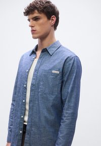 Modra srajca iz chambraya z gumbi, dolgimi rokavi, prsnim žepom in etiketo na levi strani. Gladek tekstura, sproščen kroj.