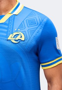 Μπλε φανέλα Los Angeles Rams με κίτρινο και λευκό γιακά, στάμπα NFL στο μανίκι και διακριτικά γεωμετρικά σχέδια στο στήθος και τον ώμο.