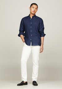 Camicia button-up di lino blu navy con maniche arrotolate, pantaloni bianchi aderenti e scarpe slip-on nere, in piedi su uno sfondo neutro.