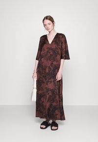 Vestido maxi em castanho com padrão floral escuro, decote em V, mangas curtas e um corte descontraído, combinado com sandálias pretas e uma mala branca.