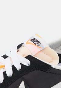 Svarta sneakers med vita snören, med en persikofärgad vadderad plös och en grå nätsektion. Märkbar Nike-logotyp på plösen.