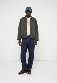Jachetă butonată verde măsliniu cu două buzunare frontale, cămașă bej dedesubt, pantaloni bleumarin și pantofi maro. Căciulă din denim cu un model în carouri.