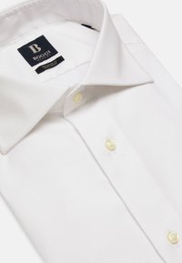 Chemise blanche à col évasé, fabriquée en tissu lisse, avec des boutons jaunes et une étiquette de logo noire sur le col.