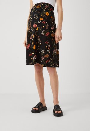 Femme portant une jupe noire fleurie longueur genou avec des fleurs orange, rose et blanches, associée à des sandales noires et un haut blanc.