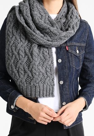 Personne portant une écharpe épaisse en maille torsadée grise par-dessus une chemise blanche et une veste en jean bleu foncé, les mains jointes à la taille.