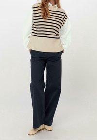 Beige a černý pruhovaný bezrukávový pletený top v kombinaci s bílými dlouhými rukávy a modrými širokými kalhotami; look doplňují béžové slip-on boty.