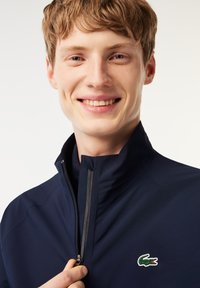 Marineblauwe zip-up jack met een hoge kraag, een gladde textuur en een groene geborduurde krokodillenlogo op de linkerborst.