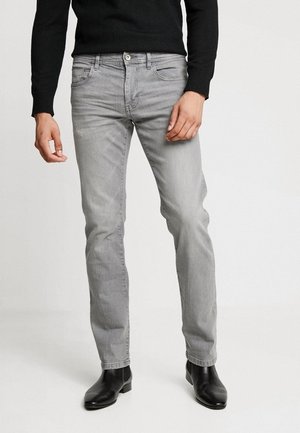 Homme portant un jean gris clair slim, un pull noir et des bottines en cuir noir, debout devant un fond clair uni.