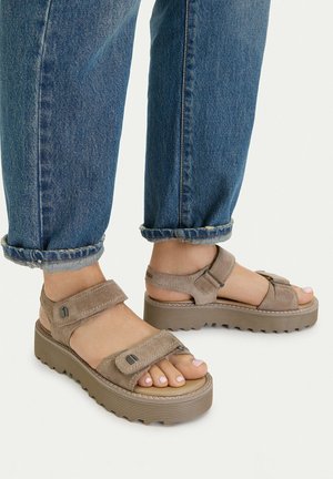 Person trägt hochgekrempelte blaue Jeans und taupefarbene Plateausandalen mit zwei verstellbaren Riemen und steht auf einer weißen Oberfläche.