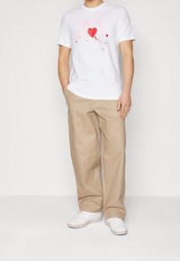 Vit bomull t-shirt med rosa och röda grafiska mönster, kombinerad med beiga vida byxor. Skorna är vita sneakers med subtila accenter.