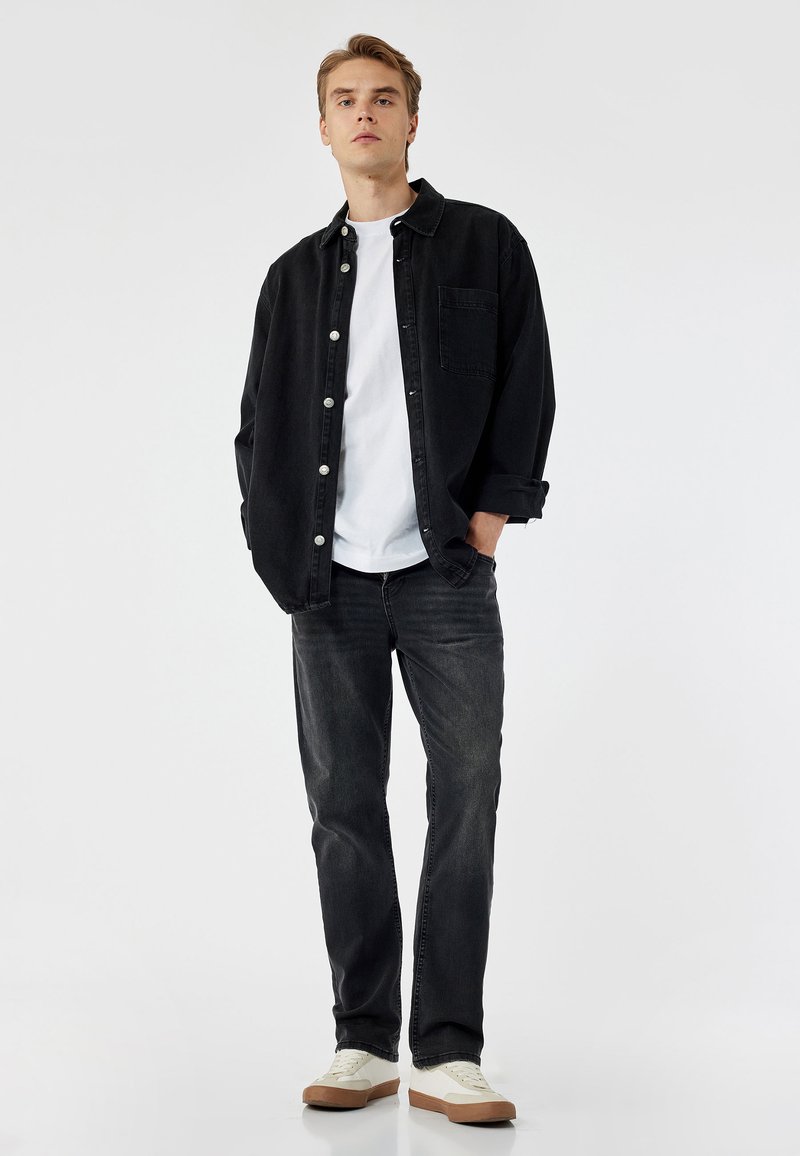 Camisa de mezclilla negra con botones blancos, llevada sobre una camiseta blanca y combinada con jeans oscuros y zapatillas blancas con suelas marrones.