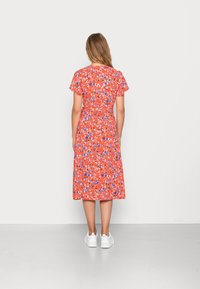 Robe portefeuille fleurie rouge avec manches courtes évasées, ornée de différentes fleurs bleues et blanches, tombant jusqu'au milieu du mollet. Portée avec des baskets blanches.