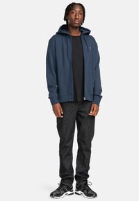 Marineblaue Kapuzenjacke mit Reißverschluss und zwei Vordertaschen, schwarzes T-Shirt darunter, dunkle Jeans und schwarze Turnschuhe mit weißen Akzenten.